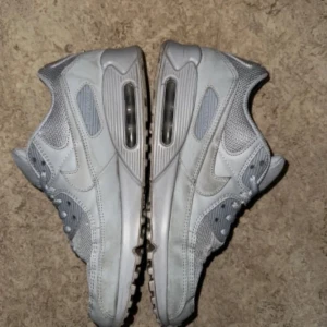 Grå Nike Air Max 90 Sneakers - Säljer ett par grå Nike Air Max sneakers i bra skick. De har den klassiska Air Max-sulan för extra komfort och stil. Perfekta för både vardag och träning. Några tecken på användning inuti, men inget som påverkar funktionen. Passar perfekt till en casual look!