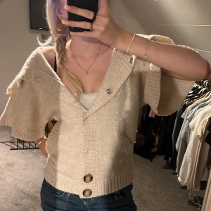 Beige stickad kofta med stora knappar - Säljer en superfin beige stickad kofta med stora knappar framtill i ullblandning. Den har en unik design med korta ärmar och en bred krage som kan bäras på olika sätt. Perfekt för både höst och vår! 🧶✨