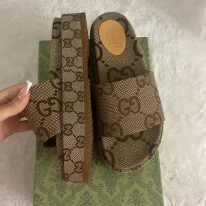 Bruna sandaler med monogram - Säljer ett par snygga bruna sandaler med monogrammönster. De har en bred rem över foten och en bekväm platt sula. Passar till både casual och mer uppklädda outfits. Box ingår