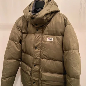 FILA Olivgrön pufferjacka  - Säljer en olivgrön pufferjacka från FILA i storlek M. Jackan är i nytt skick och har en skön, quiltad design med dragkedja och knappar. Perfekt för höst och vinter med sin varma och bekväma passform. Den har en justerbar huva och praktiska fickor. En stilren och funktionell jacka för kyliga dagar!