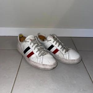 Vita sneakers från Moncler - Säljer ett par vita Moncler sneakers i okej skick. De har snygga ränder i rött och blått på sidan och klassisk snörning. Sulan är lite smutsig men annars och en rispa framtill men annars schyssta sneakers. Har varken kvitto eller original box kvar, men köpta på NK i Stockholm för 5000kr                                                                  Hör av Dig för fler bilder eller funderingar.