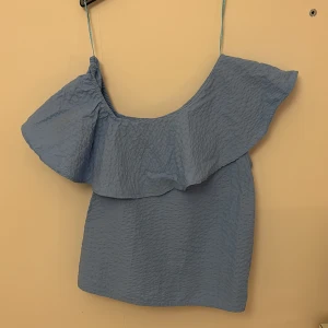 Blå offshoulder topp - Säljer en superfin blå offshoulder topp med volangdetaljer. Perfekt för sommardagar eller en festlig kväll. Toppen har en luftig passform och är tillverkad i ett lätt material som känns skönt mot huden. Den har en enkel axelbandslös design som ger en chic look💞