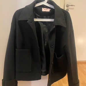 Svart kappa från Nelly - Snygg svart kappa från Nelly med knappar framtill och stora fickor. Jackan har en klassisk krage och långa ärmar med stilrena detaljer.