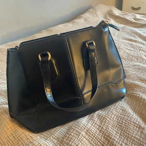 Svart axelväska med dragkedja - Snygg svart axelväska i skinn med praktisk dragkedja. Väskan har två handtag och en avtagbar axelrem för flexibilitet. Perfekt för att bära dina dagliga nödvändigheter med stil. Från Zara, finns lite metall med avskavd färg men det finns bilder på det.