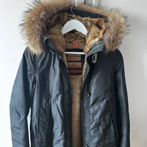 Svart parkajacka från Parajumpers - Snygg svart parkajacka från Parajumpers med pälsfodrad huva och insida. Jackan har en dragkedja och knappar framtill samt flera praktiska fickor. Perfekt för kalla vinterdagar. Det en en knapp som har gått av på jackan och det är de enda som gör att den ser använd ut! Utgångspris är 1500 men högst bud vinner 