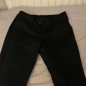 Svarta kostymbyxor - Snygga svarta kostymbyxor med låg midja, (den är lägre än vad de ser ut som på bild) och bootcut ben. Byxorna har en stilren design med knappar framtill och är perfekta för en elegant look.