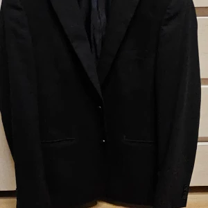 Svart kavaj från Riley - Elegant svart kavaj från Riley med klassisk design. Kavajen har en normal passform och är perfekt för formella tillfällen. Den är tillverkad i ett stilrent material och har knappar framtill.