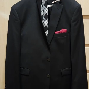 Svart kostym från Dressmann - Elegant svart kostym från Dressmann med kavaj och byxor. Kavajen har två knappar och en bröstficka med en röd näsduk. Perfekt för formella tillfällen.