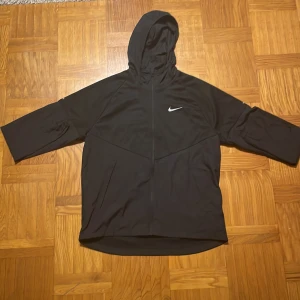 Svart vindjacka från Nike - Säljer en svart vindjacka från Nike med huva och dragkedja. Perfekt för löpning eller vardagstil med reflekterande detaljer och håller dig varm under kyliga dagar.  Vill få bort den så snabbt som möjligt!! Snabbast får!!!