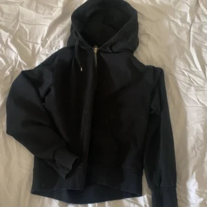 Svart hoodie med dragkedja - Säljer en klassisk svart hoodie med dragkedja och huva. Perfekt för en avslappnad stil och enkel att matcha med andra plagg. Den har långa ärmar och ribbade muddar vid ärmslut och nederkant.