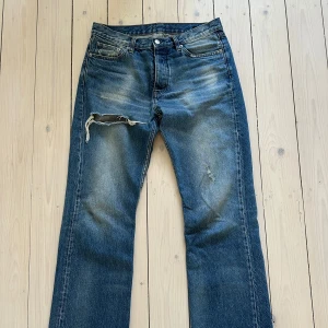 Blå jeans från HOPE modell ”rush” - Säljer ett par blå jeans ”washed” från HOPE med slitna detaljer och en klassisk femficksdesign. Jeansen har en knappgylf och är tillverkade i Portugal. Perfekta för en avslappnad stil.