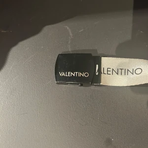 Beige bälte från Valentino - Snyggt vitt bälte från Valentino med svart spänne och logotyp. Perfekt för att ge en stilren touch till din outfit. Bältet har en modern design med tydlig branding.