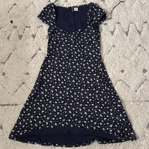 Blommig klänning från H&M - Söt blommig klänning från H&M i mörkblått med vita blommor. Klänningen har en ganska urringad hals och korta ärmar, perfekt för sommarens varma dagar. Den är tillverkad i ett lätt och bekvämt material som ger en fin passform.