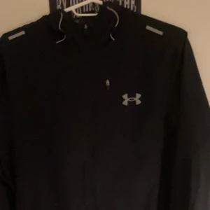 Svart vindjacka från Under Armour - Säljer en svart vindjacka från Under Armour med dragkedja och justerbar huva. Jackan har långa ärmar och reflekterande detaljer på axlarna. Perfekt för utomhusaktiviteter.