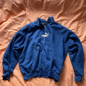 Blå tröja med dragkedja från Puma - Säljer en blå tröja från Puma med dragkedja och logotyp på bröstet. Tröjan har långa ärmar och ribbade muddar vid ärmslut och nederkant. Perfekt för en sportig look!