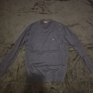 Blå tröja från Superdry - Säljer en stilren grå tröja från Superdry med rund halsringning och ett litet broderat märke på bröstet. Tröjan har långa ärmar och ribbade muddar vid ärmslut och nederkant.