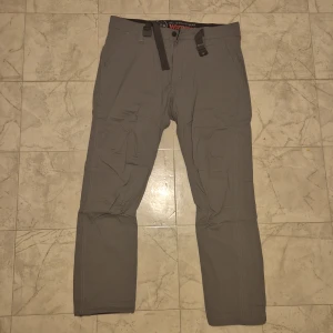 Grå byxor med bälte - Säljer ett par grå byxor med justerbart bälte och dragkedja. Byxorna har en rak passform och är perfekta för en avslappnad stil. De är tillverkade i ett slitstarkt material och har praktiska fickor både fram och bak.