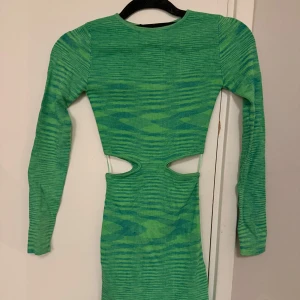 Grön långärmad klänning med cut-out detaljer -  Grön klänning med lång ärm och unika cut-out detaljer i midjan. Klänningen har ett subtilt randigt mönster som ger en extra dimension till designen. 