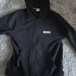 Svart hoodie från Boss - Snygg svart hoodie från Boss med dragkedja och huva. Perfekt för en avslappnad stil. Den har en enkel design med logotypen på bröstet och praktiska fickor framtill. Den är lite smutsig på bilden men tvättar den innan den säljs 