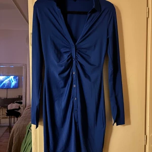 Mörkblå klänning med knappar - Elegant mörkblå klänning med långa ärmar och knappar framtill. Klänningen har en vacker rynkad detalj vid bysten som ger en smickrande passform. Perfekt för en utekväll!