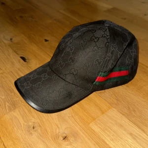 Guccikeps UNISEX - Snygg svart keps med diskret logotypmönster och klassiska gröna och röda ränder på sidan. Justerbar rem baktill för perfekt passform. Perfekt accessoar för en stilren look.
