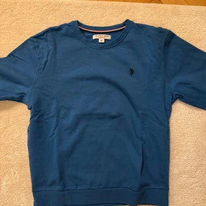 Blå tröja från U.S. Polo Assn. - Snygg blå tröja från U.S. Polo Assn. med ett diskret logotypbroderi på bröstet. Tröjan har en klassisk passform och rund halsringning. Perfekt för en avslappnad stil. Storlek M. Mycket bra skick.