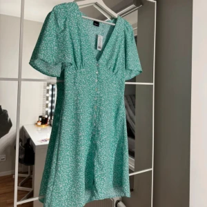 Grön blommig klänning från Gina Tricot - Söt grön klänning med vita blommor från Gina Tricot. Den har en v-ringad design med knappar framtill och korta ärmar. Perfekt för en somrig look!