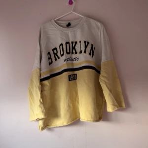 Gul och vit tröja med Brooklyn-tryck - Säljer en cool gul och vit tröja med texten 'Brooklyn athletic' och årtalet '1990' tryckt på framsidan. Tröjan har långa ärmar och en avslappnad passform. Perfekt för en casual look!