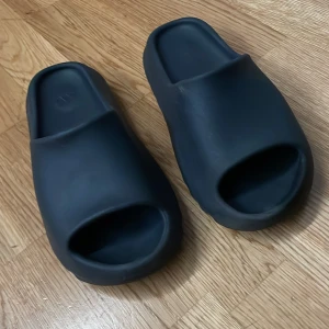 Svarta yeezy slides - Snygga svarta yeezy slides med en minimalistisk design. De har en öppen tå och en tjock sula för extra komfort. Perfekta för avslappnade dagar.