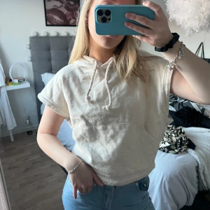 Beige kortärmad hoodie - Säljer en beige kortärmad hoodie med huva och dragsko. Perfekt för en avslappnad stil. Den har en stor ficka framtill och är gjord i ett mjukt material.