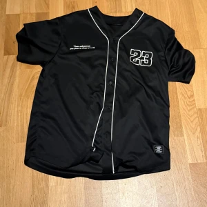 Svart baseballtröja med nummer 23 - Snygg svart baseballtröja med vita detaljer och nummer 23 på bröstet. Tröjan har knappar framtill och korta ärmar, perfekt för en sportig look.