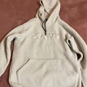 Beige fleece hoodie med ficka från märket CLWR - Mysig beige hoodie i fleece med stor ficka framtill och justerbar huva. Svarta detaljer på ärmarna ger en cool kontrast. Perfekt för kyliga dagar. Storlek SMALL men passar även medium. ord pris 900kr använt 1 gång, pris kan diskuteras.