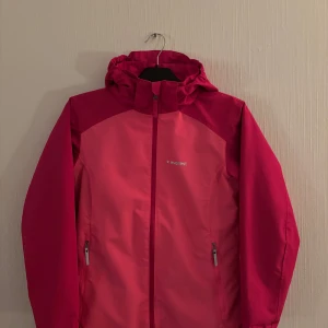 Rosa vindjacka från Everest - Snygg rosa vindjacka från Everest med dragkedja och huva som är avtagbar. Jackan har två praktiska fickor med dragkedja framtill och en reflekterande detalj på baksidan. Perfekt för blåsiga dagar. Den är justerbar i midjan och har även en innerficka! 