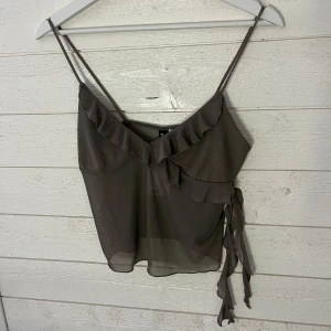 Brun linne med volangdetaljer - Snygg brun linne med smala axelband och volangdetaljer framtill. Toppen har en knytning på sidan och är perfekt för varma dagar. Den är tillverkad i ett lätt och luftigt material.