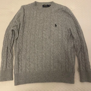 Grå stickad tröja från Polo Ralph Lauren - Säljer en stilren grå stickad tröja från Polo Ralph Lauren. Tröjan har ett klassiskt flätmönster och en broderad logotyp på bröstet. Perfekt för kyligare dagar med sin mjuka och bekväma passform. Lämna gärna prisförslag 😁