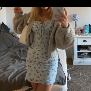 Ljusblå blommig klänning - Söt ljusblå klänning med blommigt mönster och knytning framtill. Klänningen är kort och har en figurnära passform, perfekt för sommarens varma dagar. Den har smala axelband och en feminin stil.