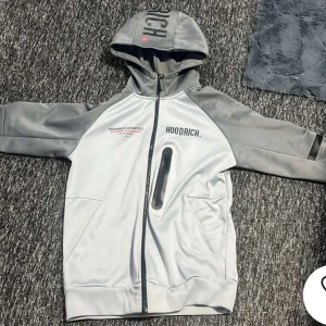 Grå och vit hoodie från Hoodrich - Säljer en snygg grå och vit hoodie från Hoodrich med textdetaljer på bröstet och ryggen. Hoodien har en dragkedja framtill och en praktisk ficka med dragkedja. Perfekt för en avslappnad stil.