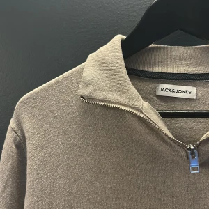 Grå tröja med dragkedja från Jack & Jones - Säljer en stilren grå tröja från Jack & Jones med dragkedja vid halsen. Perfekt för en avslappnad look. Tröjan har långa ärmar och ribbade muddar vid ärmslut och nederkant.