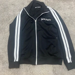 Svart träningsjacka från Palm Angels - Snygg svart träningsjacka från Palm Angels med vita ränder längs ärmarna och dragkedja framtill. Jackan har en broderad logga på bröstet och är perfekt för en sportig look. Köpt för 4680 kr äkta kofta 