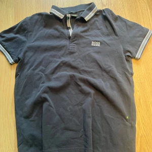 Mörkblå pikétröja från Hugo Boss - Säljer en stilren mörkblå pikétröja från Boss med klassisk krage och knappar. Tröjan har vita detaljer på kragen och ärmsluten. Perfekt för en avslappnad men ändå uppklädd look.