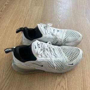 Vita Nike air 270 - Säljer ett par vita Nike Air 270 sneakers med svart detaljering och snörning. Skorna har en luftig ovandel med perforerade mönster och en synlig luftkudde i hälen för extra komfort. Perfekta för en sportig look. 