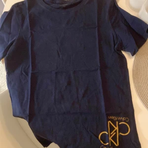 Mörkblå t-shirt från Calvin Klein - Snygg mörkblå t-shirt från Calvin Klein med deras ikoniska logga tryckt i guld på framsidan. Perfekt för en stilren och avslappnad look. Gjord i mjuk bomull för extra komfort.