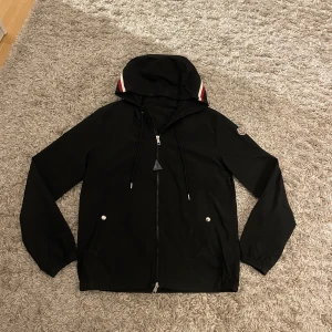 Moncler windbreaker  - Säljer en stilren svart vindjacka från Moncler med huva och dragkedja. Jackan har långa ärmar och Moncler logan på ärmen samt väldig snygg design på luvan med det röda och vita. Perfekt för dig som vill vara lite extra slitigt på dom blåsiga dagar. Var inte rädd o höra av dig!!