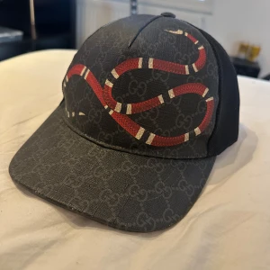 Gucci kingsnake keps - Snygg svart keps från Gucci med ett iögonfallande ormmönster i rött och vitt. Kepsen har ett justerbart band bak med röd och blå rand. Tillverkad i Italien med högkvalitativt material och stilren design. Box finns också