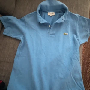 Blå pikétröja från Lacoste - Säljer en klassisk blå pikétröja från Lacoste med den ikoniska krokodilloggan på bröstet. Tröjan har korta ärmar och en bekväm passform. Perfekt för en avslappnad stil. Hör av dig vid intresse! 💙