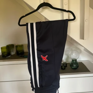 Mörkblåa träningsbyxor från Axel Arigato - Snygga mötkblåa träningsbyxor från Axel Arigato med vita ränder längs sidorna och en röd logotyp. Byxorna har elastisk midja, byxorna är marin blå. Köpt i Arigaro butik i Göteborg och är knappt använda