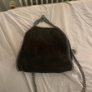 Svart axelväska med kedjedetaljer - Snygg svart axelväska med coola kedjedetaljer runt kanterna och som axelrem. Väskan har en stilren design och är perfekt för att ge din outfit en edgy touch. Tillverkad i ett material som liknar mocka.