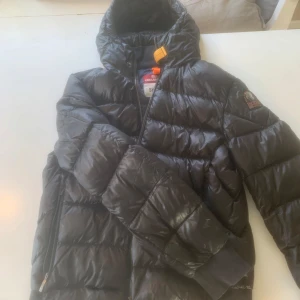 Svart dunjacka från Parajumpers - Säljer en svart dunjacka från Parajumpers med huva och dragkedja. Jackan har en quiltad design och är perfekt för kyligare väder. Den har långa ärmar och en snygg logga på ärmen. Perfekt för dig som vill hålla stilen i vinter! Köpt på Johnells för cirka 7000kr