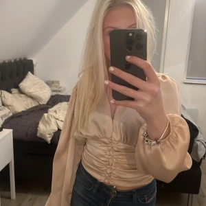 Beige omlottblus - Säljer en elegant beige omlottblus med långa ärmar och v-ringning. Blusen har en smickrande rynkad detalj framtill som ger en fin passform. Perfekt för en stilren look.
