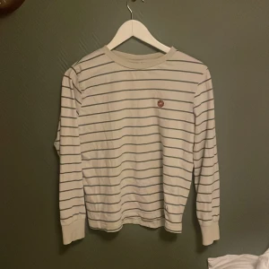 Vit och blå randig tröja från Grunt - Säljer en stilren vit tröja med gråa ränder från Grunt. Tröjan har långa ärmar och en liten röd detalj på bröstet. Perfekt för en avslappnad stil. Strl L motsvarar ungefär 14 år ( 158/164) 😁